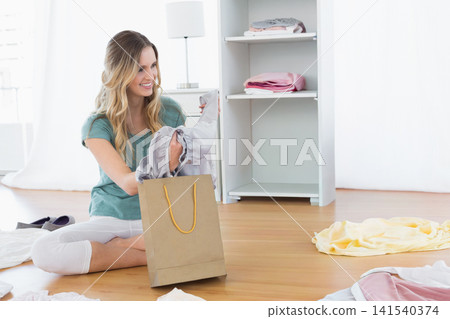 Woman sitting on bedroom floor holding gray blouse unpacking from brown bag with yellow handles Woman sitting on bedroom floor holding gray blouse unpacking from brown bag with yellow handles 141540374