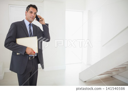 Mature man standing left in modern entry wearing suit and tie holding smartphone portfolio with pen Mature man standing left in modern entry wearing suit and tie holding smartphone portfolio with pen 141540386