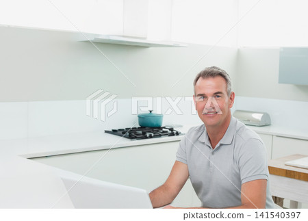 Mature man sitting at kitchen peninsula using laptop beside teal Dutch oven and toaster, copy space Mature man sitting at kitchen peninsula using laptop beside teal Dutch oven and toaster, copy space 141540397