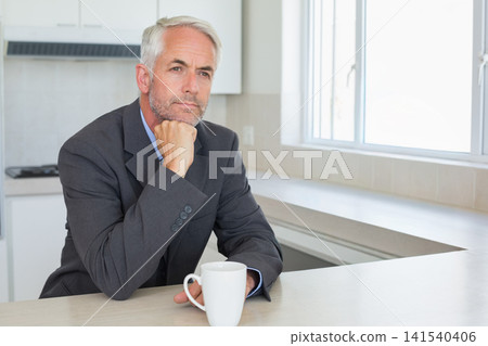 Mature man sitting at kitchen counter holding white mug, resting chin, wearing dark jacket Mature man sitting at kitchen counter holding white mug, resting chin, wearing dark jacket 141540406