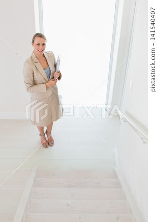 Professional woman in beige skirt suit standing on foyer landing holding clipboard and tote Professional woman in beige skirt suit standing on foyer landing holding clipboard and tote 141540407