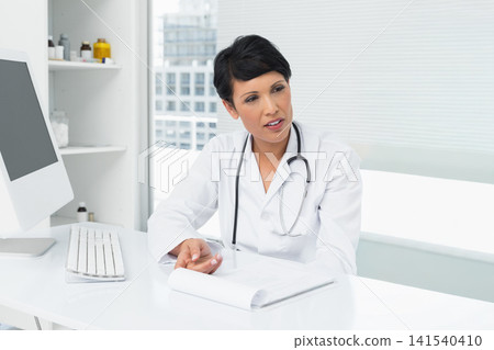 Female physician sitting at white desk in clinic, wearing white lab coat and stethoscope, gesturing Female physician sitting at white desk in clinic, wearing white lab coat and stethoscope, gesturing 141540410