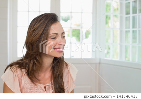 Mid-adult woman sitting in sunroom near grid windows with white wall and sill smiling looking right Mid-adult woman sitting in sunroom near grid windows with white wall and sill smiling looking right 141540415