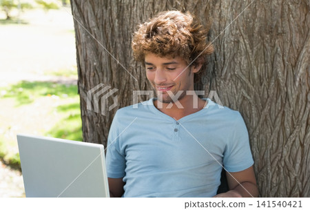 Man sitting against textured tree trunk in park, using silver laptop and showing wristband Man sitting against textured tree trunk in park, using silver laptop and showing wristband 141540421