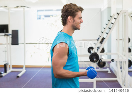 Man wearing sleeveless turquoise shirt holding blue dumbbell and training in gym with weight rack Man wearing sleeveless turquoise shirt holding blue dumbbell and training in gym with weight rack 141540426