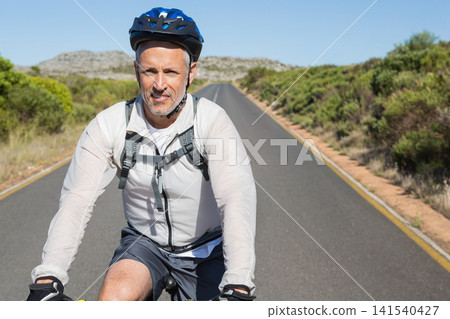 Mature man riding bicycle toward camera on paved road wearing blue helmet and hydration pack Mature man riding bicycle toward camera on paved road wearing blue helmet and hydration pack 141540427