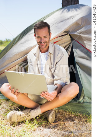 Mature man sitting at tent entrance wearing beige shirt and khaki shorts using laptop 141540429