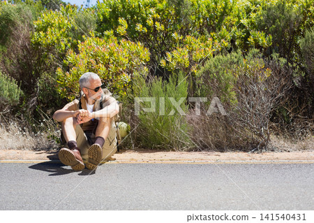 Middle-aged man sitting on paved shoulder beside shrubs wearing sunglasses, shorts, backpack, boots Middle-aged man sitting on paved shoulder beside shrubs wearing sunglasses, shorts, backpack, boots 141540431