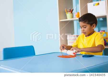 Asian 5yo boy yellow-shirt sitting at blue-table gluing red-tip glue to orange paper, copy space Asian 5yo boy yellow-shirt sitting at blue-table gluing red-tip glue to orange paper, copy space 141540435