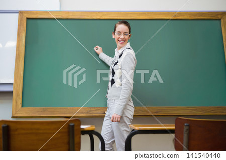 Female teacher standing front of class holding chalk at chalkboard, argyle sweater, glasses, desks Female teacher standing front of class holding chalk at chalkboard, argyle sweater, glasses, desks 141540440