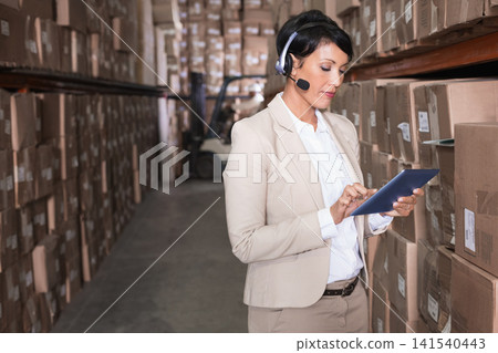 Mature adult woman wearing beige suit using tablet and headset in warehouse aisle with forklift 141540443