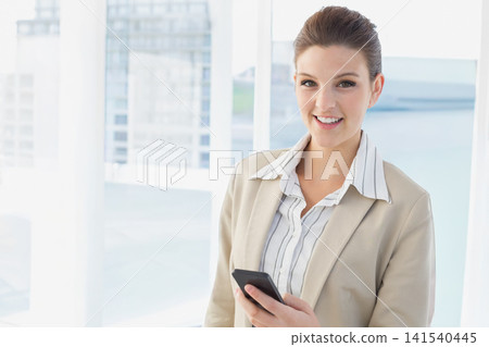 Female office worker wearing beige blazer standing by tall windows holding phone, copy space Female office worker wearing beige blazer standing by tall windows holding phone, copy space 141540445