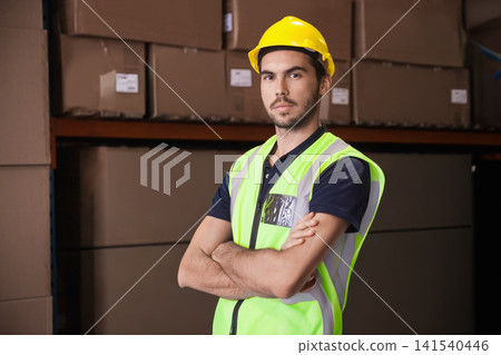 Man standing arms crossed by warehouse shelf with cartons, wearing yellow hard hat and safety vest Man standing arms crossed by warehouse shelf with cartons, wearing yellow hard hat and safety vest 141540446