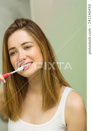 Woman brushing teeth at bathroom sink holding pink toothbrush and wearing white tank top Woman brushing teeth at bathroom sink holding pink toothbrush and wearing white tank top 141540450
