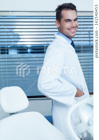 Male dentist standing on right side of dental clinic with chair wearing white lab coat, smiling Male dentist standing on right side of dental clinic with chair wearing white lab coat, smiling 141540453