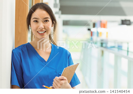 Asian female nurse leaning on post in clinic hall in blue scrubs holding clipboard yellow pencil Asian female nurse leaning on post in clinic hall in blue scrubs holding clipboard yellow pencil 141540454