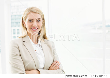 Female manager standing with crossed arms by tall windows, beige blazer, city view, copy space 141540455