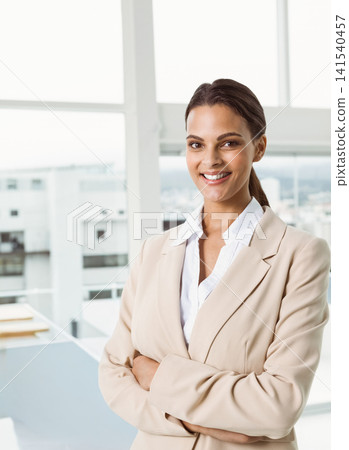 Woman standing by large window in office with stacked folders, smiling in beige blazer, white shirt Woman standing by large window in office with stacked folders, smiling in beige blazer, white shirt 141540457