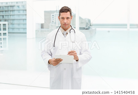 Male physician standing in clinic holding tablet and pen, wearing lab coat and stethoscope Male physician standing in clinic holding tablet and pen, wearing lab coat and stethoscope 141540458