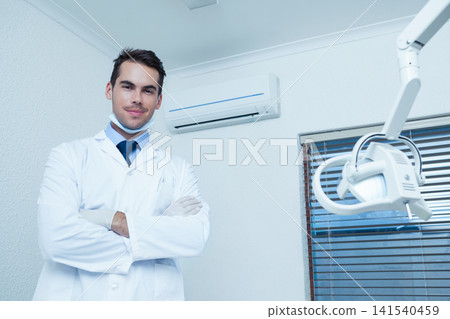 Male dentist standing arms crossed in clinic wearing white lab coat near dental lamp, copy space Male dentist standing arms crossed in clinic wearing white lab coat near dental lamp, copy space 141540459