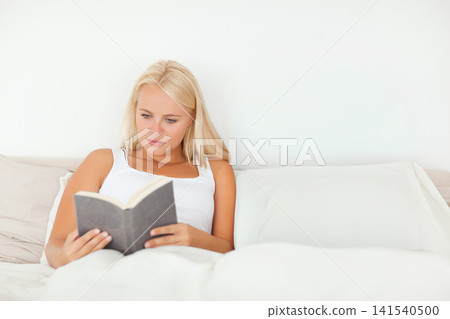 Blonde woman reclining on bed at home, reading paperback book in soft white tank top Blonde woman reclining on bed at home, reading paperback book in soft white tank top 141540500
