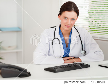 Female physician sitting at clinic consultation desk leaning forward with stethoscope and keyboard Female physician sitting at clinic consultation desk leaning forward with stethoscope and keyboard 141540501