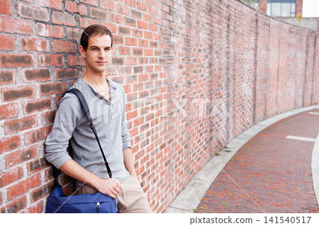 Man leaning against red brick wall on walkway, in henley, carrying blue messenger bag, copy space Man leaning against red brick wall on walkway, in henley, carrying blue messenger bag, copy space 141540517