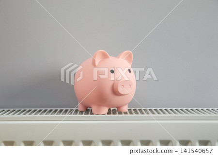 Heating season. Piggy bank sits on top of a radiator during heating season Heating season. Piggy bank sits on top of a radiator during heating season 141540657