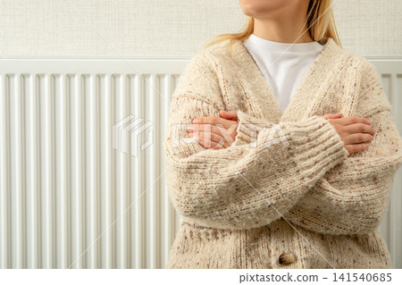 Heating season. Woman in warm sweater stands by radiator, staying warm during heating season 141540685
