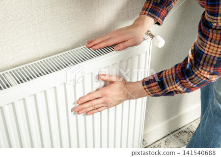 Heating season. Person checks warmth of radiator in cozy home environment. Heating season concept 141540688