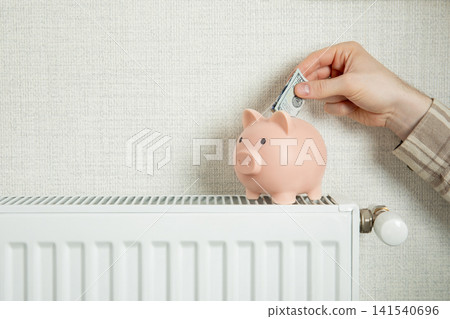 Heating season. Hand inserts cash into piggy bank placed on radiator Heating season. Hand inserts cash into piggy bank placed on radiator 141540696