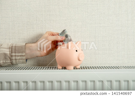 Heating season. Hand places money into piggy bank sitting on home radiator Heating season. Hand places money into piggy bank sitting on home radiator 141540697