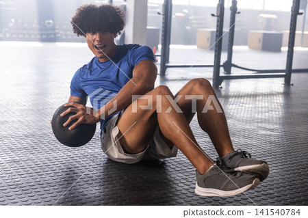African American man in sportswear twisting torso and holding black medicine ball on gym floor African American man in sportswear twisting torso and holding black medicine ball on gym floor 141540784