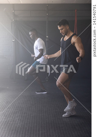 Diverse male friends in sportswear skipping jump ropes on rubber mat in gym beside squat rack Diverse male friends in sportswear skipping jump ropes on rubber mat in gym beside squat rack 141540791