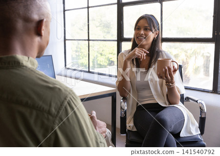 Diverse coworkers talking at office by window holding coffee mug near laptop, copy space Diverse coworkers talking at office by window holding coffee mug near laptop, copy space 141540792