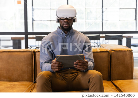 African american man sitting on office lounge sofa wearing white vr headset, holding tablet African american man sitting on office lounge sofa wearing white vr headset, holding tablet 141540800