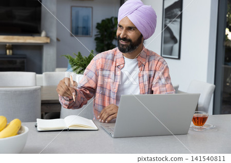 Mid adult indian man wearing lavender turban writing in notebook at countertop table with laptop Mid adult indian man wearing lavender turban writing in notebook at countertop table with laptop 141540811