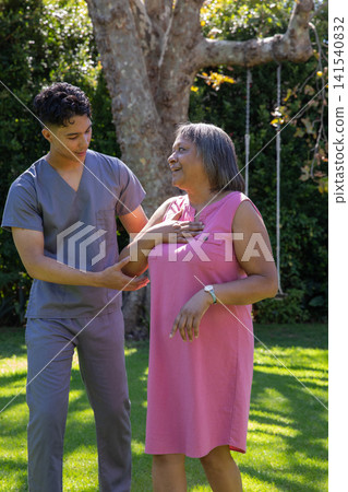 Caregiver wearing scrubs supporting senior woman standing on lawn under tree with rope swing 141540832