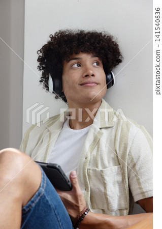 African american male sitting on floor at home wearing headphones, holding smartphone African american male sitting on floor at home wearing headphones, holding smartphone 141540836