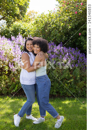 Mother and daughter hugging and smiling on lawn in backyard garden with purple shrubs Mother and daughter hugging and smiling on lawn in backyard garden with purple shrubs 141540853