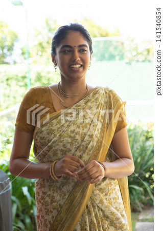 Indian woman standing on sunlit garden patio, in floral sari mustard-patterned and gold jewelry Indian woman standing on sunlit garden patio, in floral sari mustard-patterned and gold jewelry 141540854