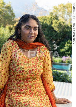 Indian woman sitting on bench in garden terrace, wearing yellow kurta with orange dupatta by pool Indian woman sitting on bench in garden terrace, wearing yellow kurta with orange dupatta by pool 141540858