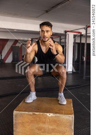 Athletic man squatting on wooden plyometric box in gym near battle ropes, extending arms forward Athletic man squatting on wooden plyometric box in gym near battle ropes, extending arms forward 141540872