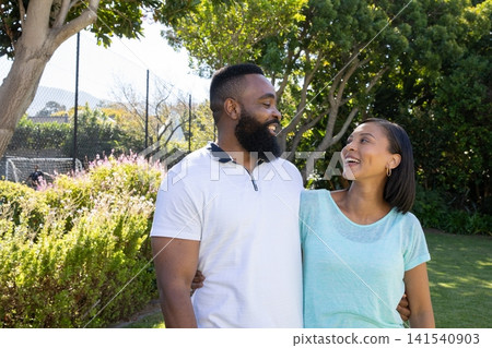 Diverse couple standing in sunlit park hugging at waist by chain link fence near flowering shrubs Diverse couple standing in sunlit park hugging at waist by chain link fence near flowering shrubs 141540903