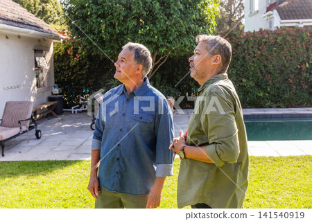 Senior male friends standing on backyard lawn beside pool and lounge chairs looking upward Senior male friends standing on backyard lawn beside pool and lounge chairs looking upward 141540919