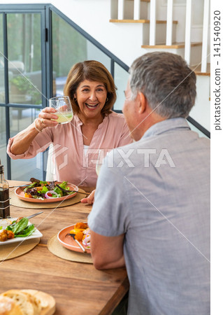 Couple sitting in dining room with woman raising pale drink glass beside salad on table 141540920