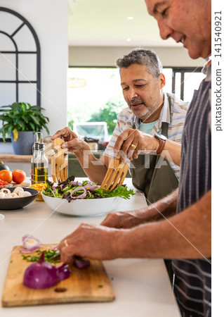 Senior male friends wearing aprons tossing salad with servers and chopping onions on kitchen island Senior male friends wearing aprons tossing salad with servers and chopping onions on kitchen island 141540921