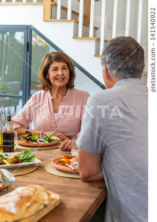 Senior couple chatting at dining table in home with salad dish, bread loaf, oil bottles Senior couple chatting at dining table in home with salad dish, bread loaf, oil bottles 141540922