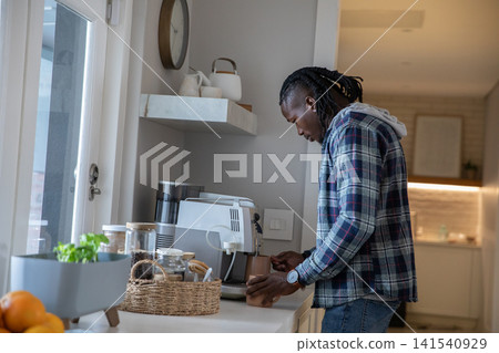 African american man placing ceramic cup under espresso machine on kitchen countertop African american man placing ceramic cup under espresso machine on kitchen countertop 141540929
