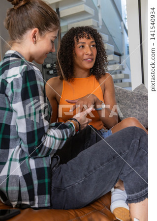 Diverse female friends sitting on couch in living room showing ring and holding smartphone Diverse female friends sitting on couch in living room showing ring and holding smartphone 141540934